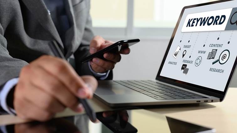 Muž v obleku drží mobil a pero u notebooku, na jehož obrazovce je schéma analýzy klíčových slov Keyword s ikonami SEO procesu.
